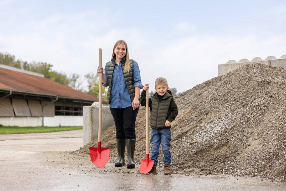 Frankfurter Schaufel mit Stiel Mini für Kinder, ideal für Gartenarbeiten, neben Erwachsenenschaufel vor Kieshaufen präsentiert.