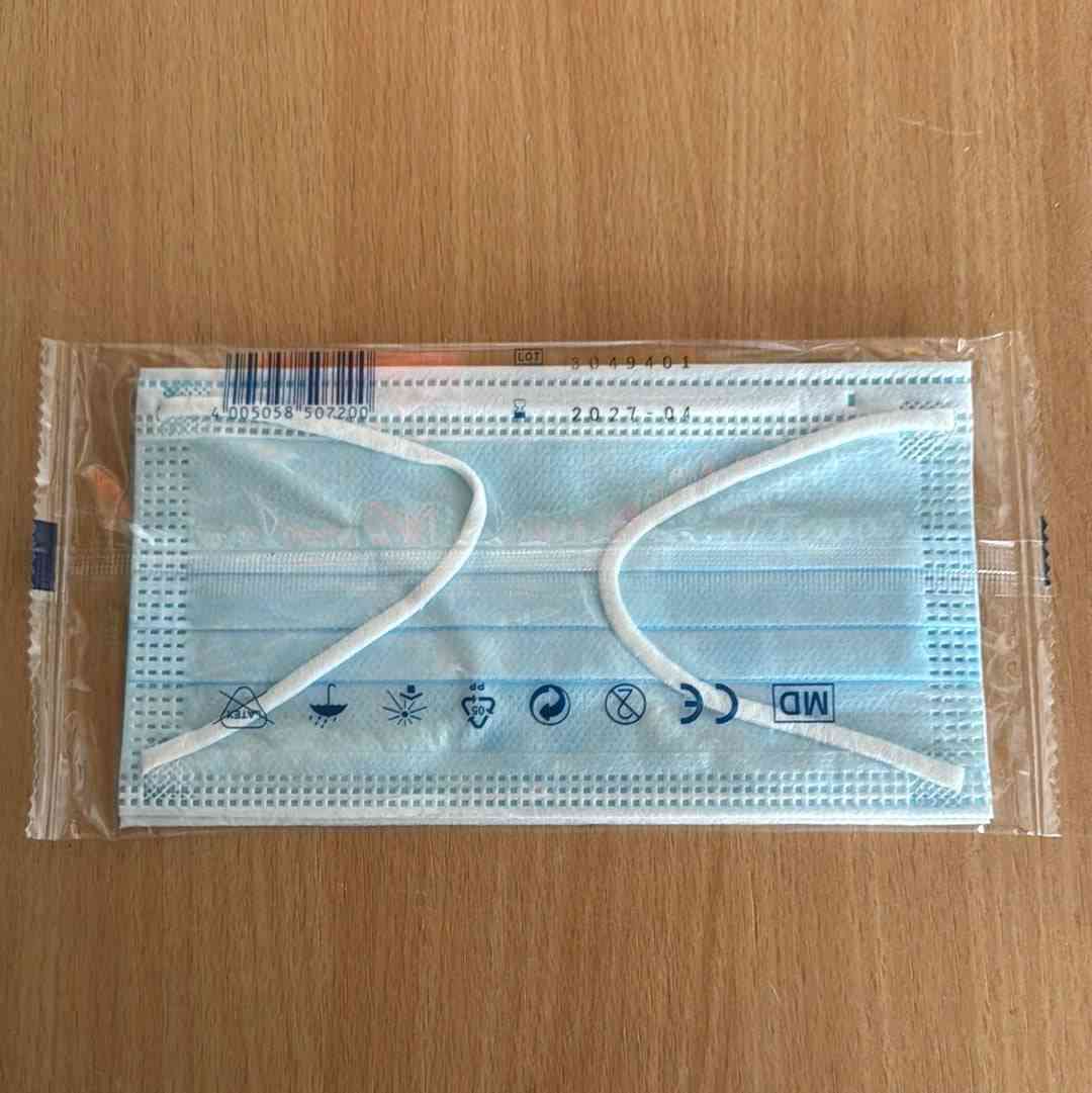 Einweg-Gesichtsmaske in transparenter Kunststoffverpackung auf Holzoberfläche, mit sichtbaren Symbolen für medizinische Verwendung, CE-Kennzeichnung und Haltbarkeitsdatum.