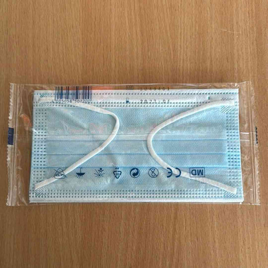 Einweg-Gesichtsmaske in transparenter Kunststoffverpackung auf Holzoberfläche, mit sichtbaren Symbolen für medizinische Verwendung, CE-Kennzeichnung und Haltbarkeitsdatum.